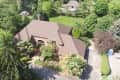 Aerial view of a large brick house with a brown roof, surrounded by lush green trees and a driveway.