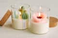 Two glass candles with cactus and flower designs, lit on a textured surface, with cork lids beside them.