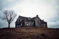 Old, weathered house on a hill with a bare tree under a cloudy sky.