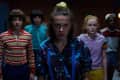 Five teenagers in colorful 80s outfits stand in a dimly lit room with brick walls.