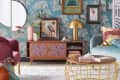 Eclectic living room with teal palm wallpaper, colorful art, pink and teal furniture, and a gold lamp on a wooden console.