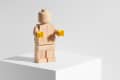 Wooden block figure with yellow hands on a white pedestal.
