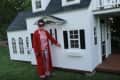 Woman in red pajamas standing beside a white playhouse with black shutters and a porch.