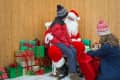 Child sitting on Santa's lap with another child handing a gift, surrounded by colorful wrapped presents.