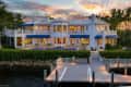 Waterfront mansion with blue awnings, large windows, and a dock, surrounded by palm trees at sunset.