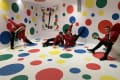 Four people in red jackets playing Twister in a room with colorful dots on walls and floor.