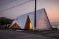 A-frame house with white siding and large windows, illuminated from inside at dusk, located on a quiet street.