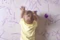 Child in yellow shirt drawing with purple marker on wall covered in scribbles.
