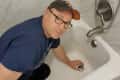 Man in a cap and glasses kneeling in a bathtub, adjusting the drain stopper.