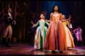 Performers in period costumes on stage, with a woman in an orange dress leading, surrounded by others in colorful gowns.