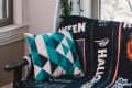 Teal geometric pillow on a chair with a Halloween-themed blanket, near a window with a hanging plant.