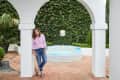 Woman in a pink shirt and jeans leaning against a white archway near a small round pool and lush greenery.