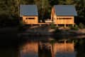 Two wooden cabins with metal roofs by a lake, surrounded by trees, with a person relaxing on a deck.