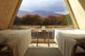 A-frame cabin interior with two beds, a small table, and a large triangular window overlooking autumn trees and mountains.