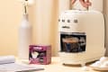 Smeg espresso machine with a cup of coffee, Lavazza pods, and a vase with flowers on a beige table.