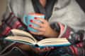 Person wrapped in a blanket holding a blue mug, reading a book.