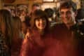 Two people smiling in a crowded bar, with warm lighting and blurred background.