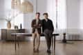 Two men standing in a bright room with wooden floors, wicker pendant lights, and a dark wooden table.