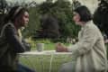 Two women sitting at a white metal table in a park, engaged in conversation, with trees in the background.