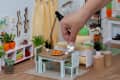 Hand placing a tiny teapot on a miniature dining table with chairs, surrounded by small plants and decor in a dollhouse setting.