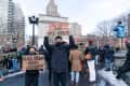 Protesters holding signs for unity and solidarity in a city park with an arch and buildings in the background.