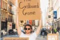 Man holding a cardboard sign reading "The Cardboard Game" on a busy city street.