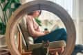 Woman reading on a chair inside a large circular cat wheel, with a tabby cat sitting in the wheel.