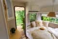 Cozy camper interior with a bed, fluffy pillows, straw hat, and a view of lush greenery through the open door.