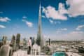 A person in a white shirt and cap stands on a rooftop, overlooking the Burj Khalifa and Dubai skyline under a blue sky.