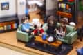 Four LEGO figures sitting on a green sofa in a living room, with a coffee table and bookshelf in the background.