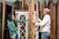 Two people examining a vintage wooden door in a workshop filled with reclaimed doors.