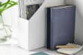 Books and magazines in a white holder, with a blue book, notepad, and pen on a desk.