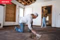 Man inspecting floor damage in a room with boarded windows, woman observing.