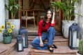Woman in red shirt and jeans sitting on terracotta steps, surrounded by lanterns, sunflowers, and potted plants.