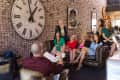 Group of people in a modern office with a large wall clock and brick walls, sitting on leather sofas and chairs.