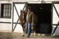 Man in cowboy hat leading a horse out of a white barn with black trim.