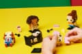 Hands assembling block figures on a yellow table with colorful background.