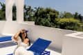 Woman in a white dress sitting on a blue lounge chair on a sunny terrace with trees in the background.
