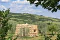 Wooden geometric structure in a lush landscape with olive trees and distant wind turbines under a blue sky.