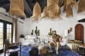 Living room with wicker pendant lights, white sofa, cactus decor, and a man sitting on a wooden chair, blue patterned rug.