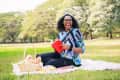 Woman sitting on a picnic blanket in a park, holding a red book and smiling, with a basket nearby.
