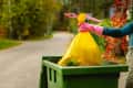 Person wearing pink gloves placing a yellow trash bag into a green bin on a tree-lined street.