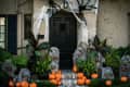 Front porch with Halloween decor, including pumpkins, tombstones, spider webs, and lush green plants.