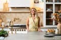 Woman in a yellow dress standing in a modern kitchen with gold fixtures, pears on the counter, and decorative plants.