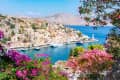 Colorful houses along a harbor with boats, surrounded by vibrant flowers and hills under a clear blue sky.
