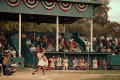 Baseball game with a female batter in a vintage uniform, catcher, umpire, and spectators in a green wooden stadium.