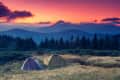 Two tents on a grassy field with a forest and mountains at sunset.