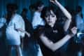 Young woman in a black dress dancing at a dimly lit party with people in white outfits.