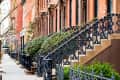 Row of brownstone buildings with ornate black iron railings and greenery along the sidewalk.