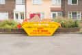 Yellow skip bin filled with debris in front of brick houses and flowering bushes.